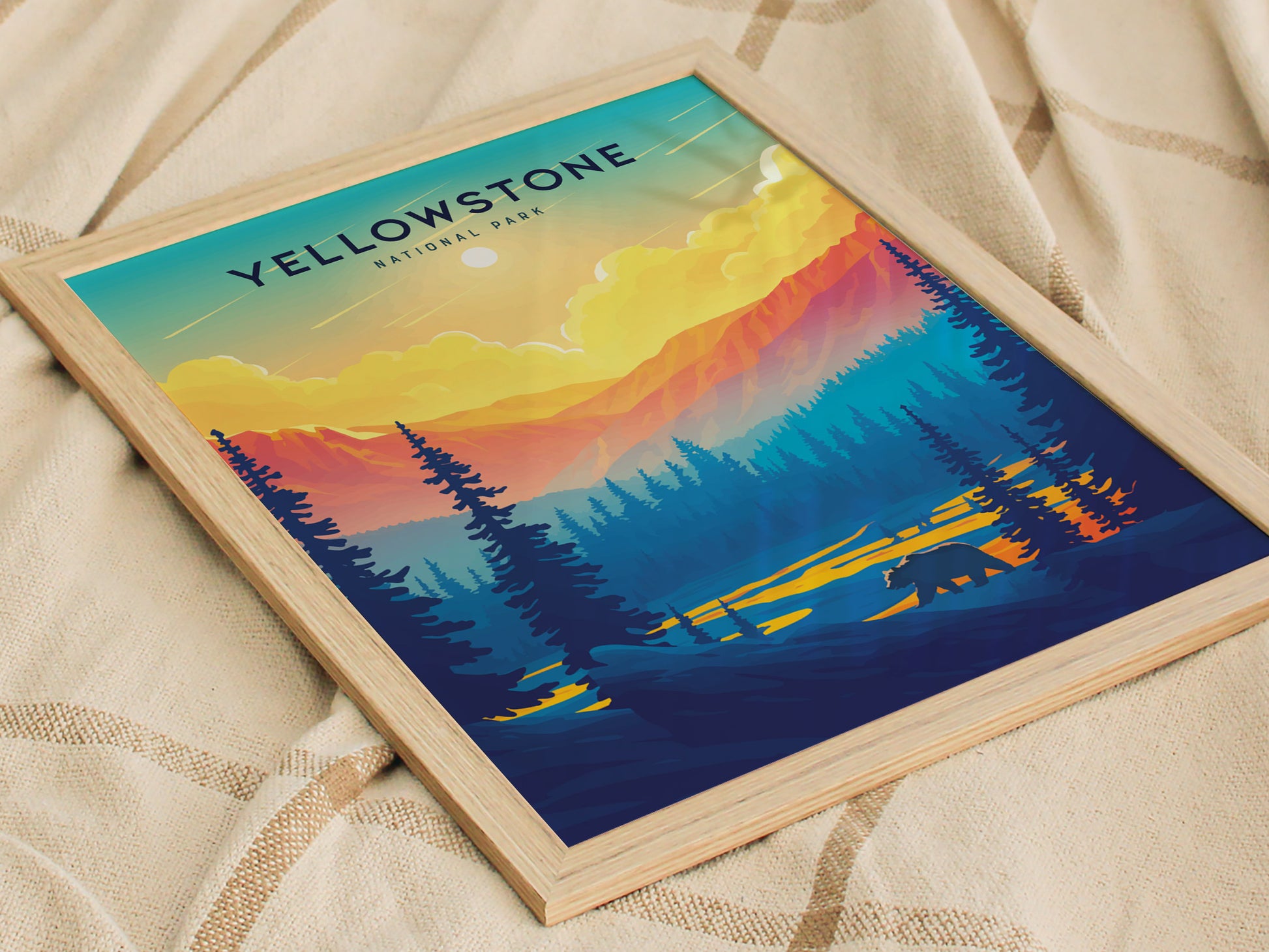 A framed poster depicting a vibrant sunset over Yellowstone National Park, featuring colorful mountains, silhouetted trees, and a bear. The artwork rests on a textured beige fabric.