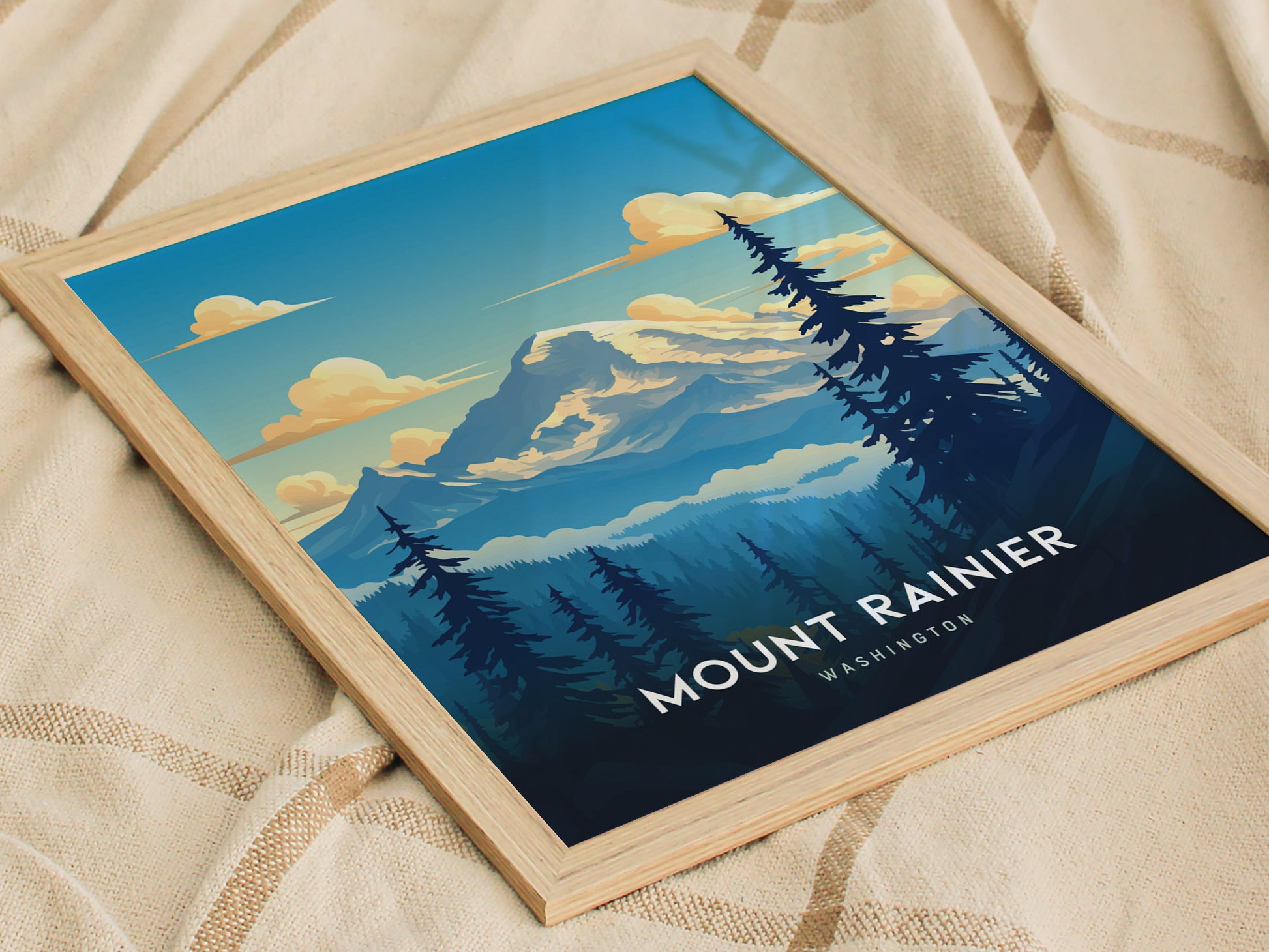 Framed poster of Mount Rainier with a stylized depiction of the mountain, surrounded by evergreen trees and a clear blue sky, rests on a beige textured fabric.