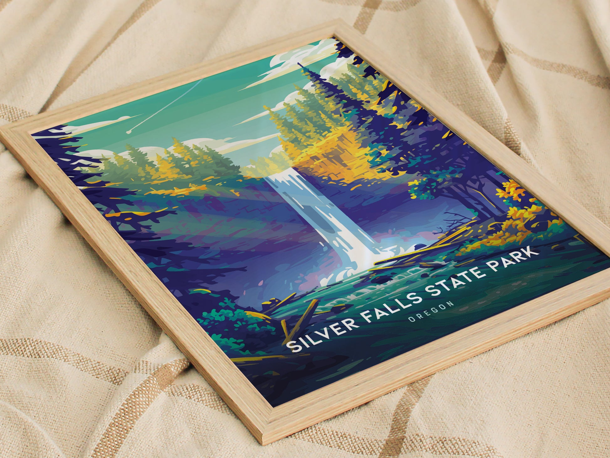 A framed poster of Silver Falls State Park features a stylized waterfall surrounded by vibrant trees under a teal sky. Its placed on a beige fabric surface.