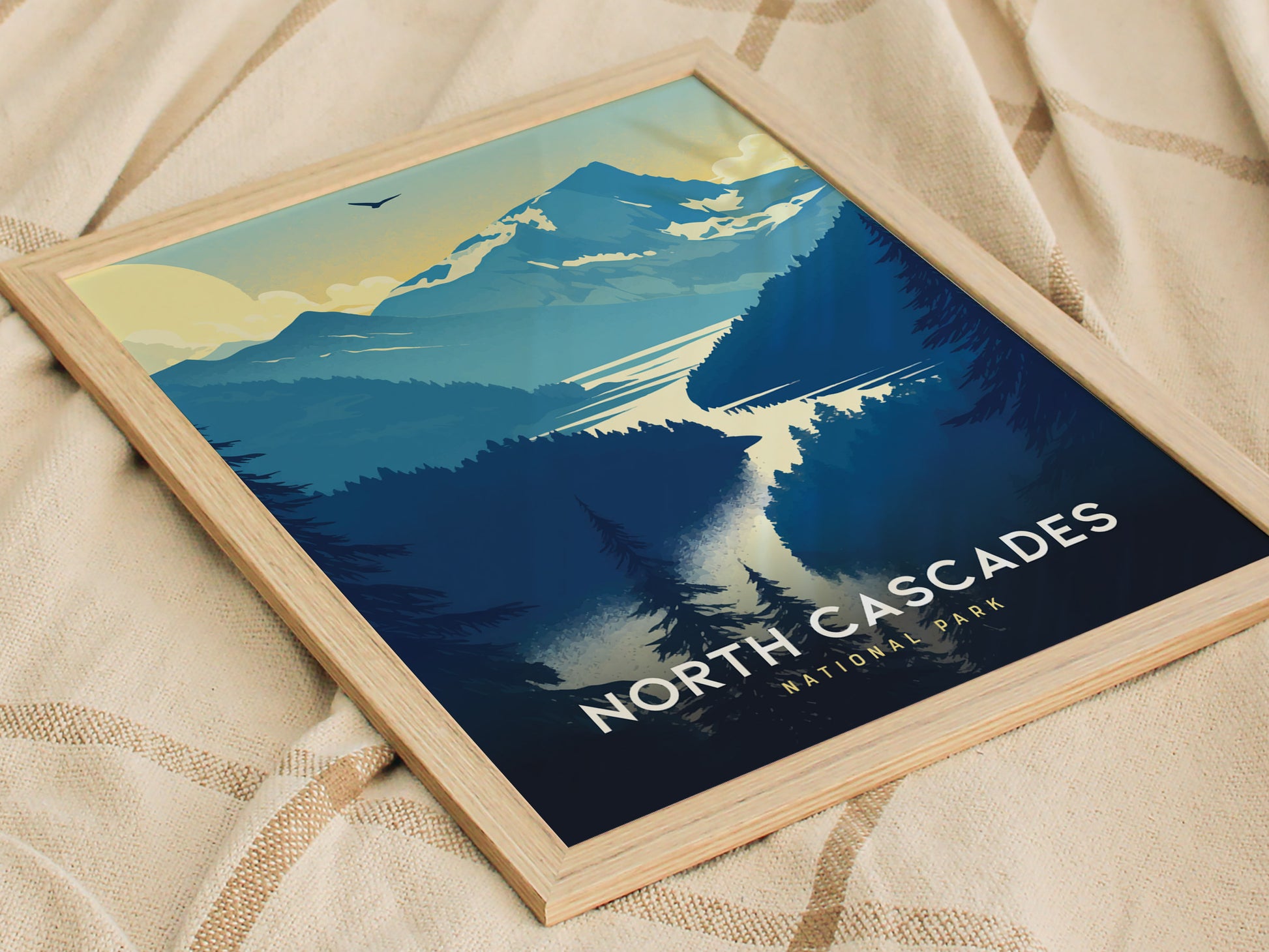 A framed poster of North Cascades National Park features stylized blue and yellow mountains, a winding river, and forest silhouettes, set against a soft fabric background.