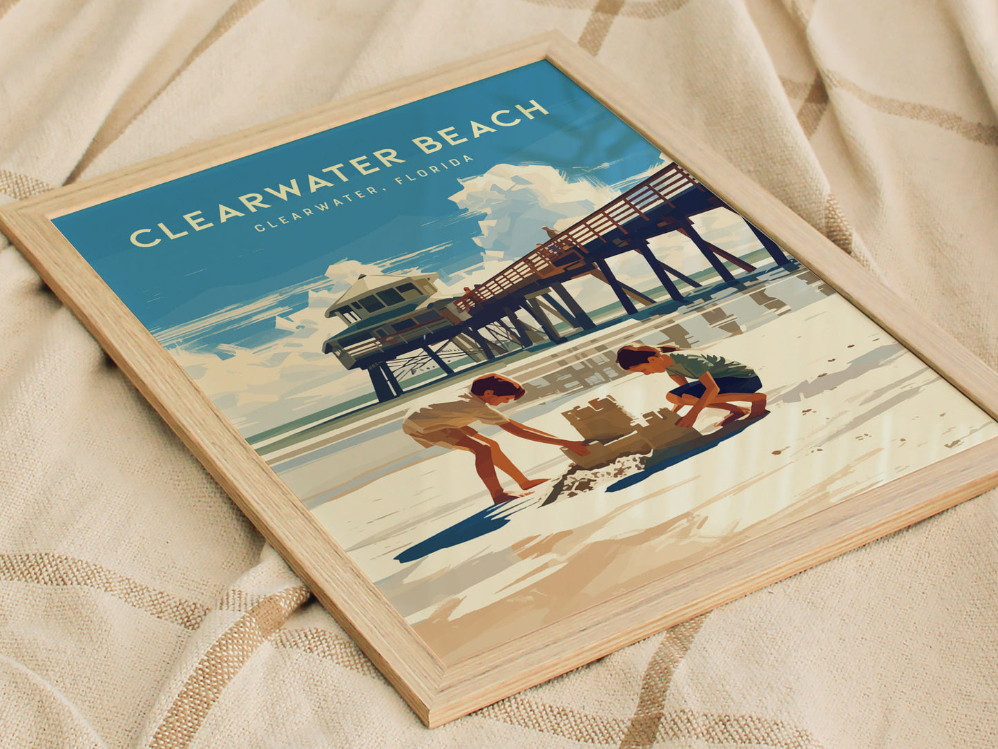 A framed poster of Clearwater Beach, Florida, shows two children building a sandcastle near a pier under a sunny sky, displayed on a textured fabric surface.