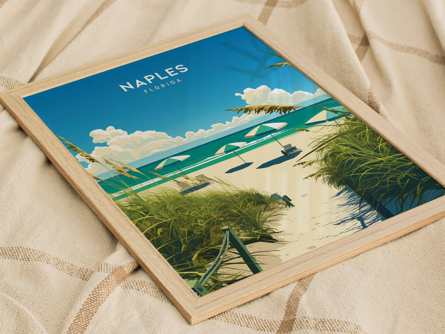 Framed artwork depicting a beach scene with sandy path, green foliage, umbrellas, and a clear blue sky, placed on a textured fabric background. Text reads Naples Florida.