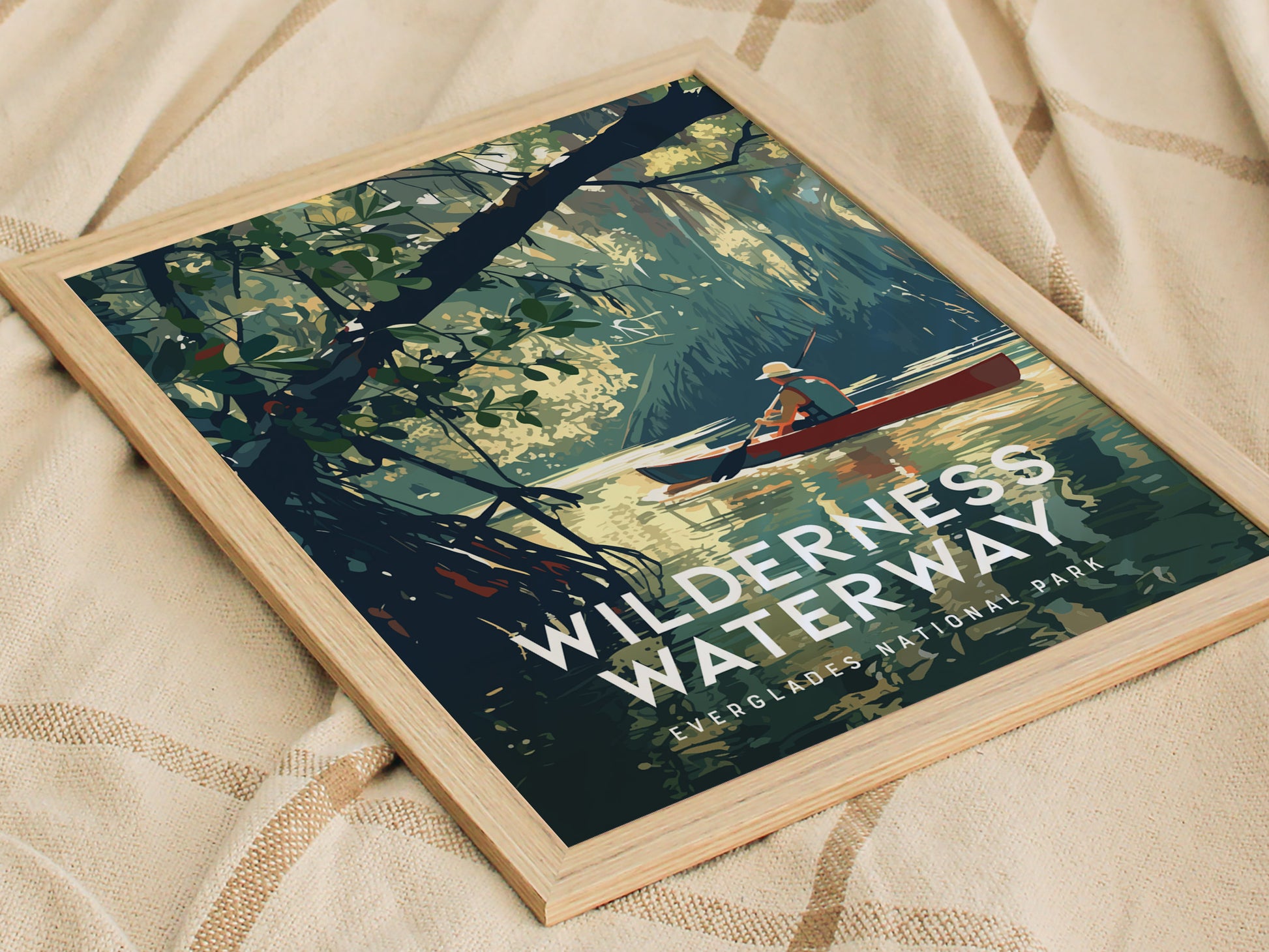 A framed poster titled Wilderness Waterway depicts a person canoeing through a serene waterway surrounded by lush mangrove forests, resting on a textured fabric surface.