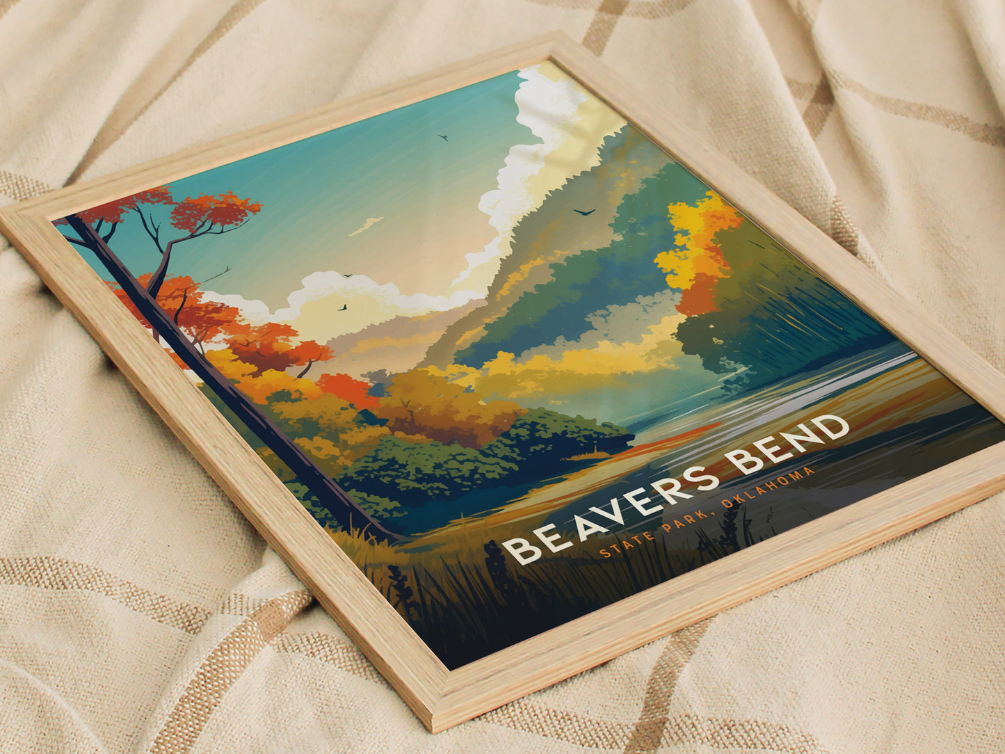 Framed poster of Beavers Bend State Park displaying colorful autumn trees, mountains, and a river under a vibrant sky. The artwork rests on a textured beige fabric.