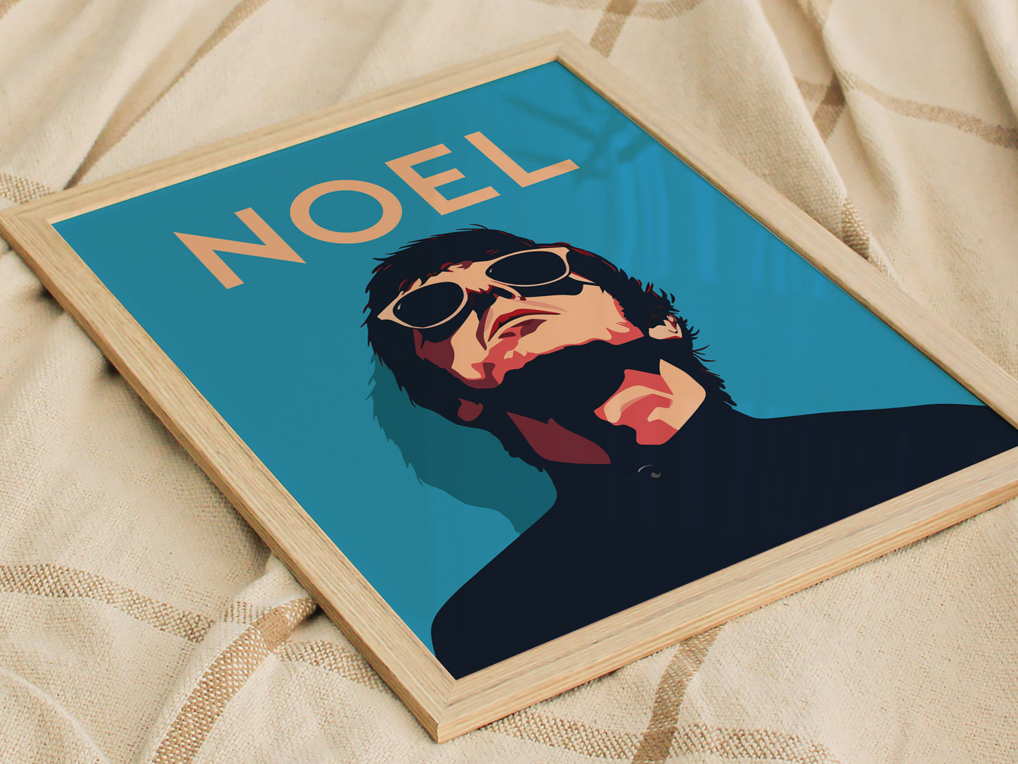 Framed poster of a man in sunglasses against a blue background with the word NOEL at the top, placed on a beige fabric surface.