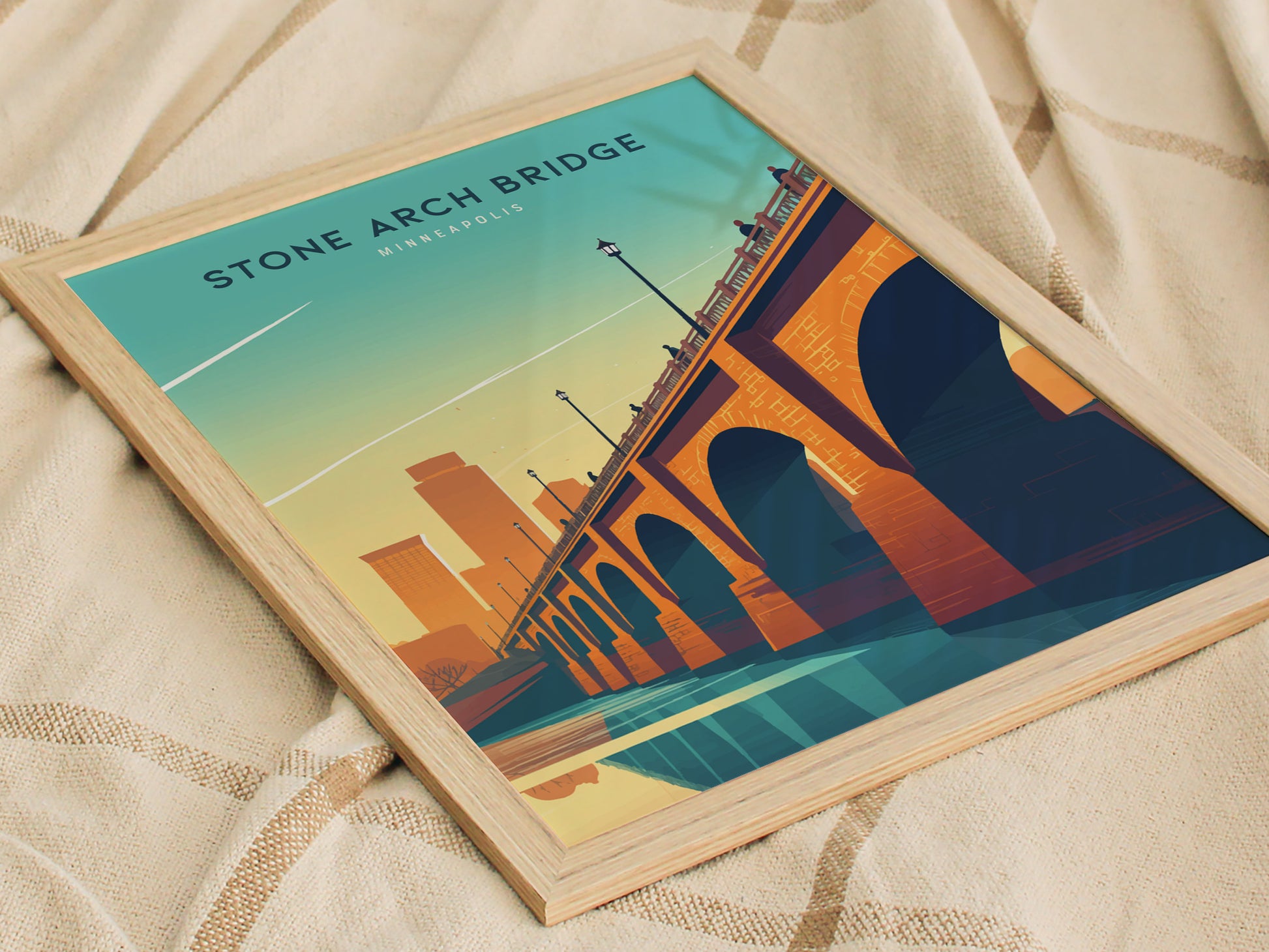 A framed poster of the Stone Arch Bridge with a stylized Minneapolis skyline is placed on a beige textured blanket. The artwork features vibrant orange.