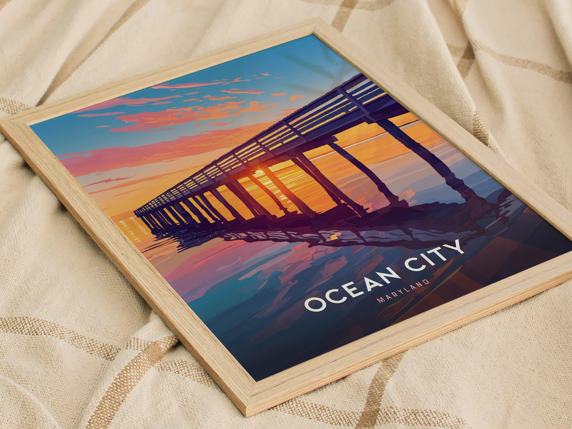 A framed poster featuring a vibrant sunset view of a boardwalk pier at Ocean City, Maryland, with colorful reflections on the water, resting on a beige fabric surface.