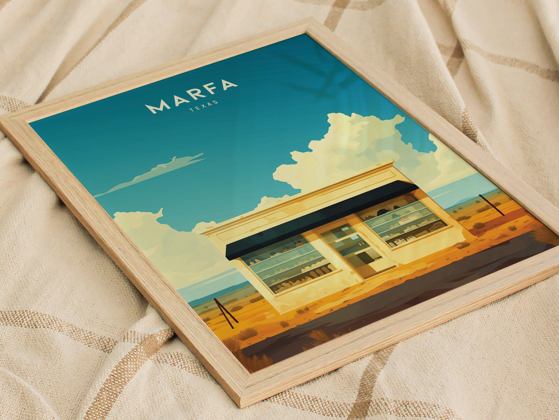 Framed Marfa, Texas poster featuring a minimalist desert scene with a small building under a vibrant blue sky, resting on a textured fabric surface.