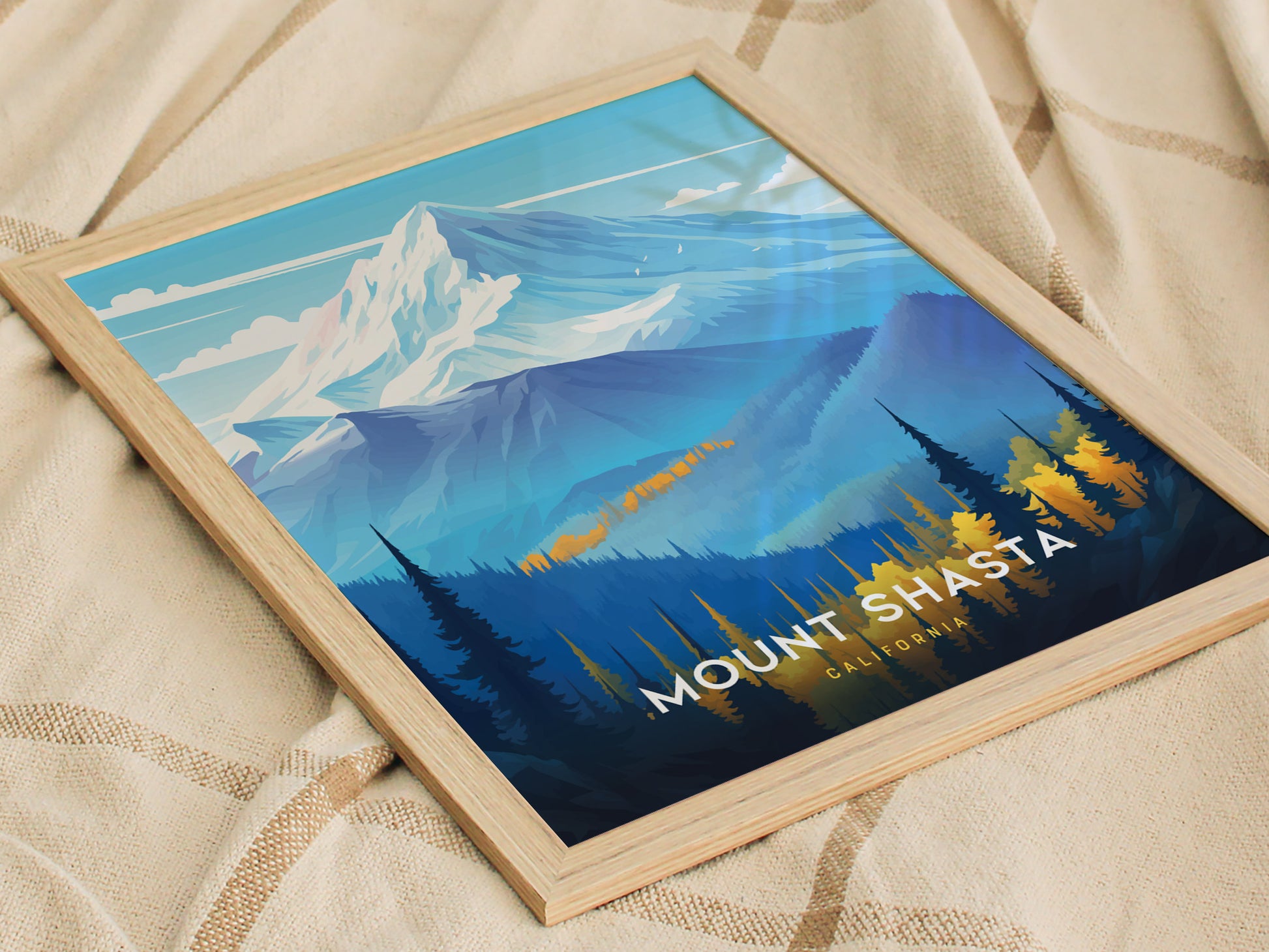 A framed poster of Mount Shasta featuring stylized snow-capped peaks and forested foothills, resting on a beige textured blanket. The text reads Mount Shasta, California.