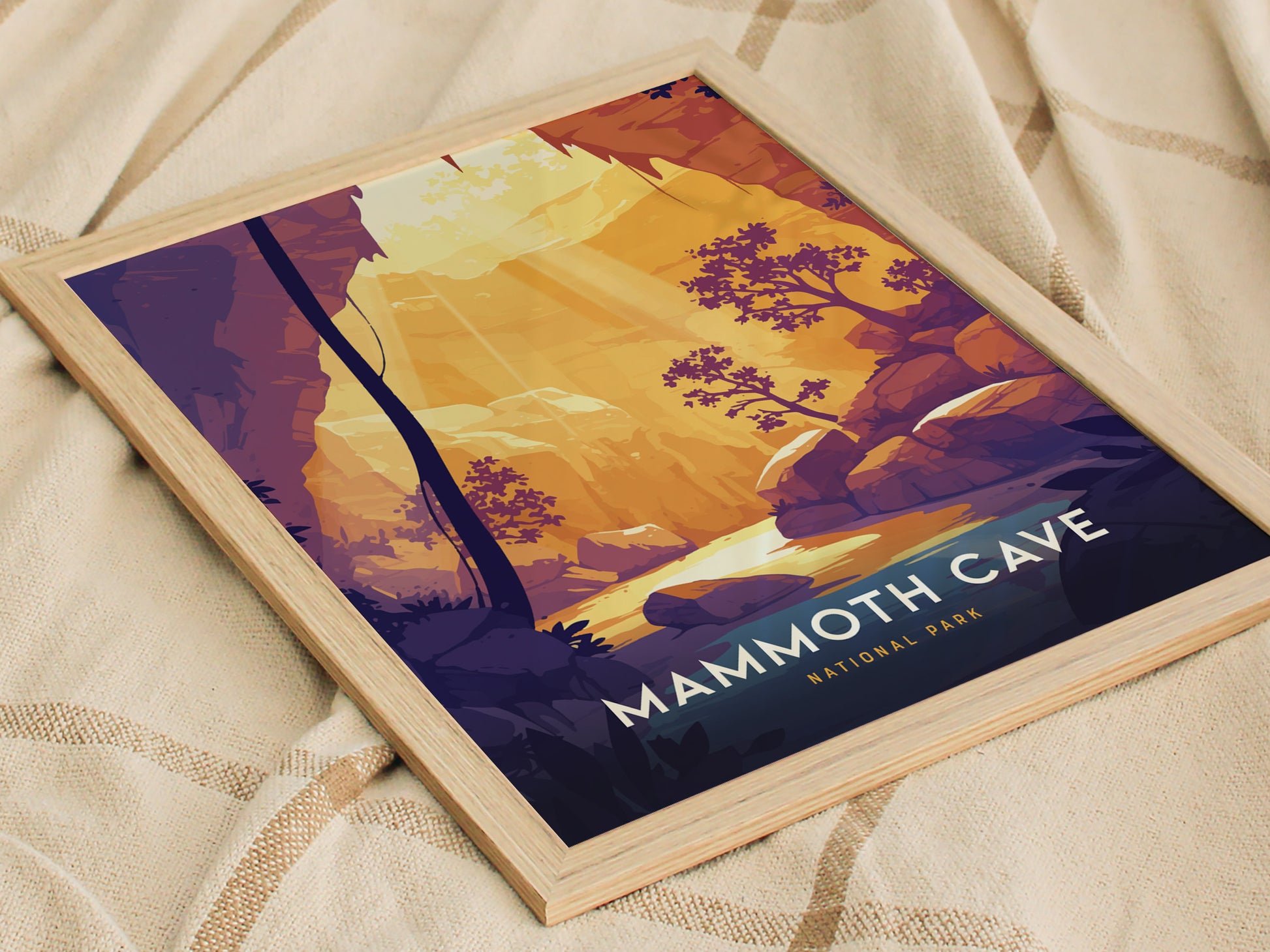 A framed poster of Mammoth Cave National Park featuring stylized illustrations of trees and rock formations in warm tones, resting on a textured fabric surface.