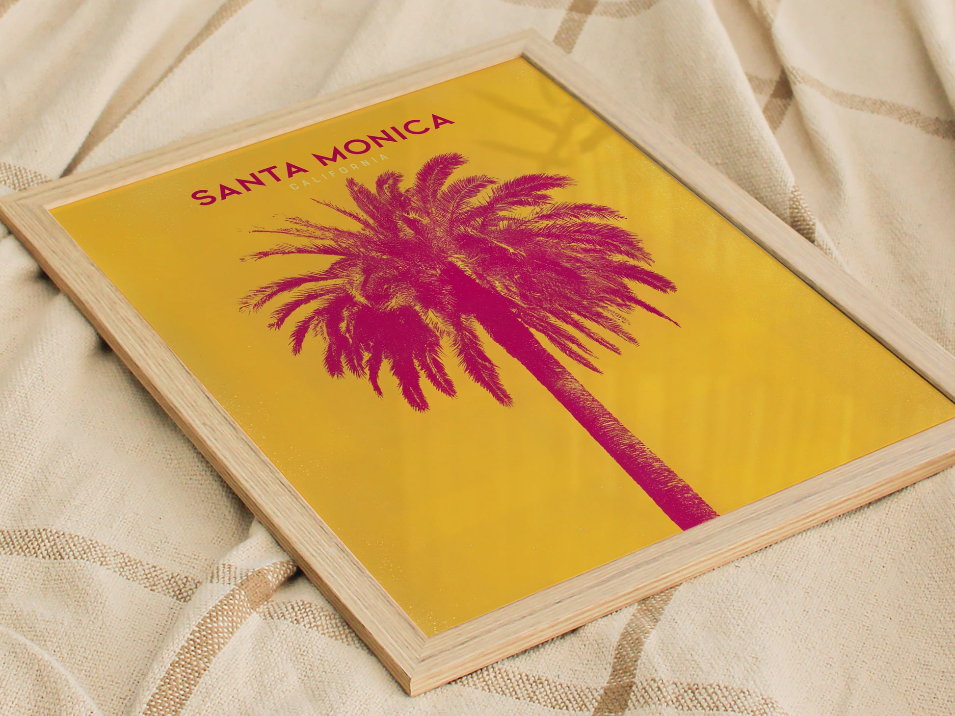 A framed poster with a vibrant pink palm tree against a yellow background, titled Santa Monica California, is placed on a textured fabric surface.