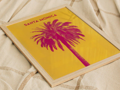 A framed poster with a vibrant pink palm tree against a yellow background, titled Santa Monica California, is placed on a textured fabric surface.