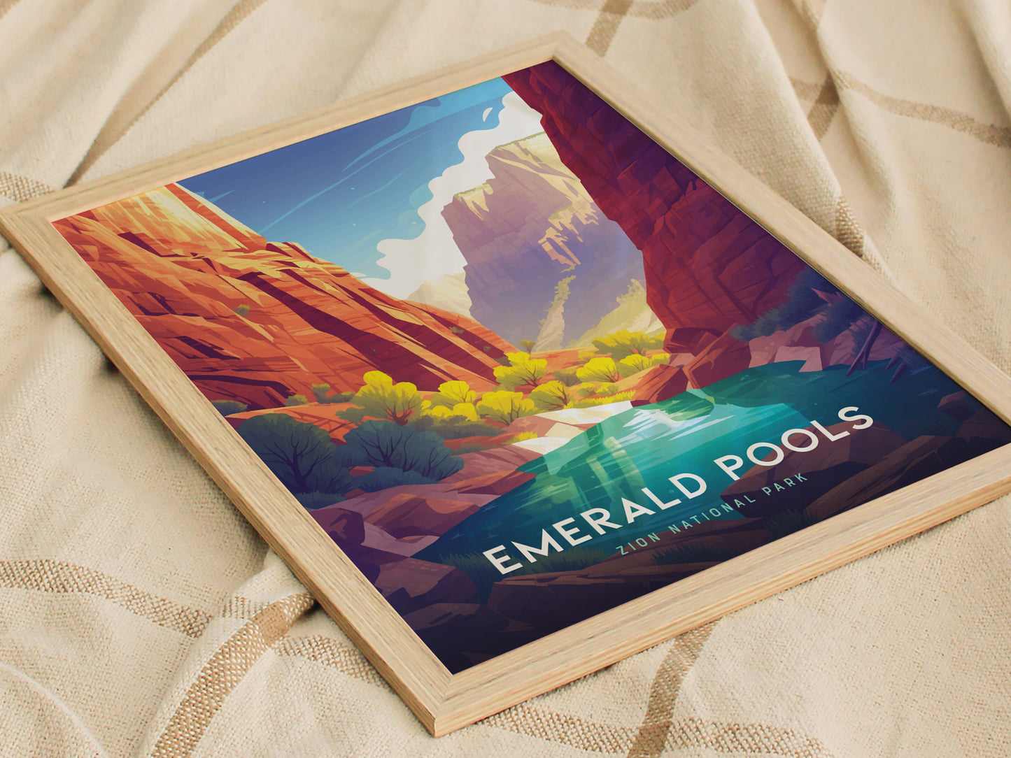A framed poster of Zion National Parks Emerald Pools, featuring vibrant red canyon cliffs and lush greenery, rests on a beige fabric surface. The artwork is colorful and inviting.