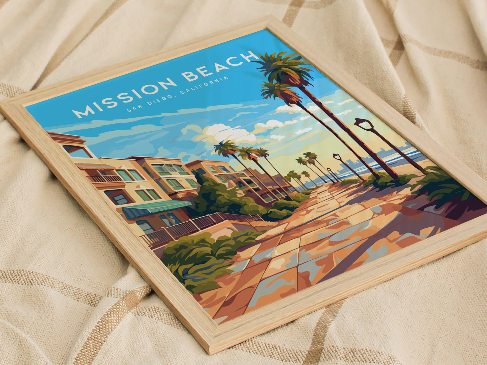 Framed artwork depicting a vibrant boardwalk scene at Mission Beach, San Diego, with palm trees, buildings, and a beach view under a blue sky, resting on a fabric background.