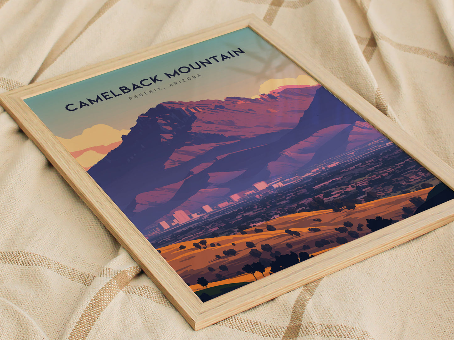 A framed poster of Camelback Mountain features a stylized landscape with rich purple and orange tones, set against a light blue sky. It rests on a beige fabric.