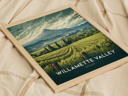 A framed illustration of Willamette Valley, Oregon, featuring rolling vineyard hills and a mountainous backdrop, rests on a textured fabric surface. The artwork highlights lush greenery and serene landscapes.