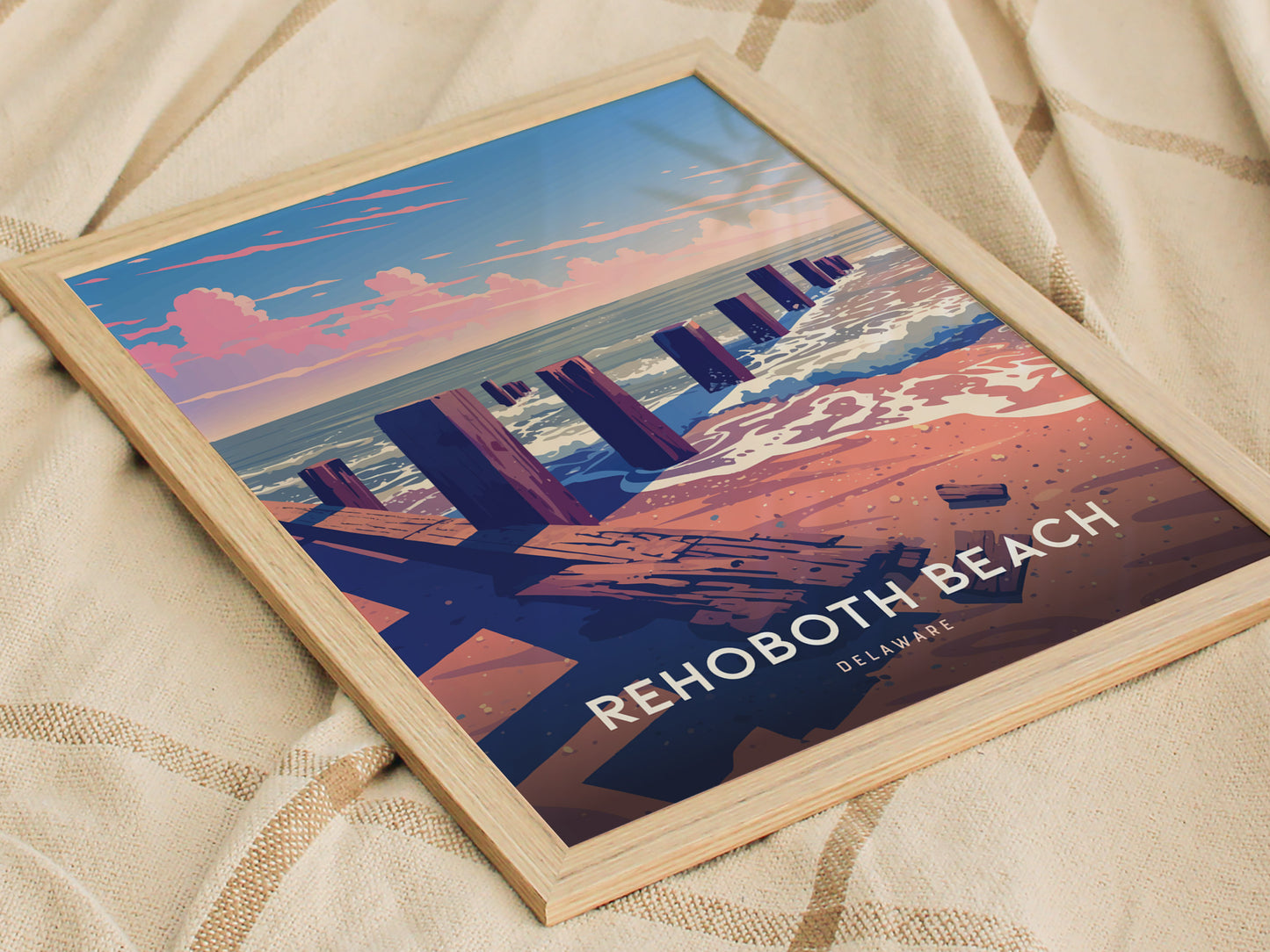 A framed poster of Rehoboth Beach, Delaware, featuring a stylized boardwalk and ocean scene with pink clouds and waves, rests on a textured beige fabric.