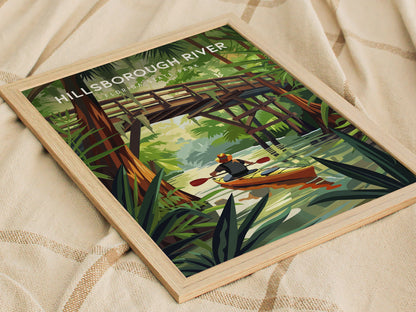 A framed poster depicting a kayaker paddling beneath a wooden bridge on a lush river, surrounded by cypress trees and foliage, is laid on a beige textured fabric.