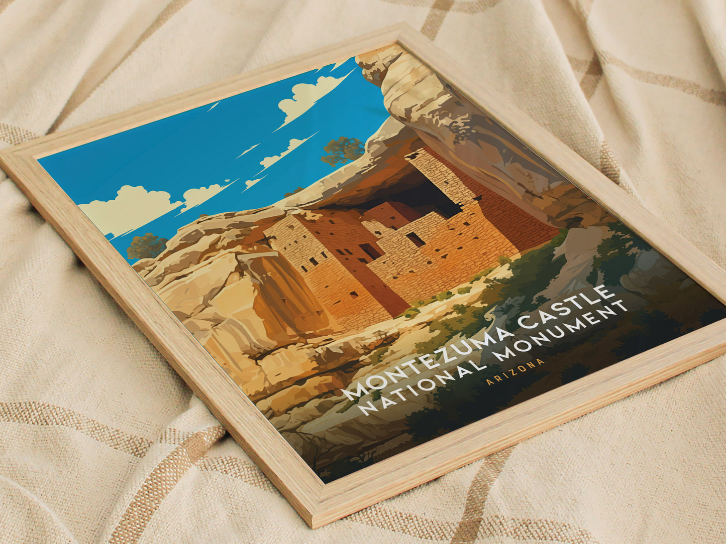 Framed poster of Montezuma Castle National Monument featuring a stylized illustration of the cliff dwelling against a vibrant blue sky, resting on a textured beige fabric.