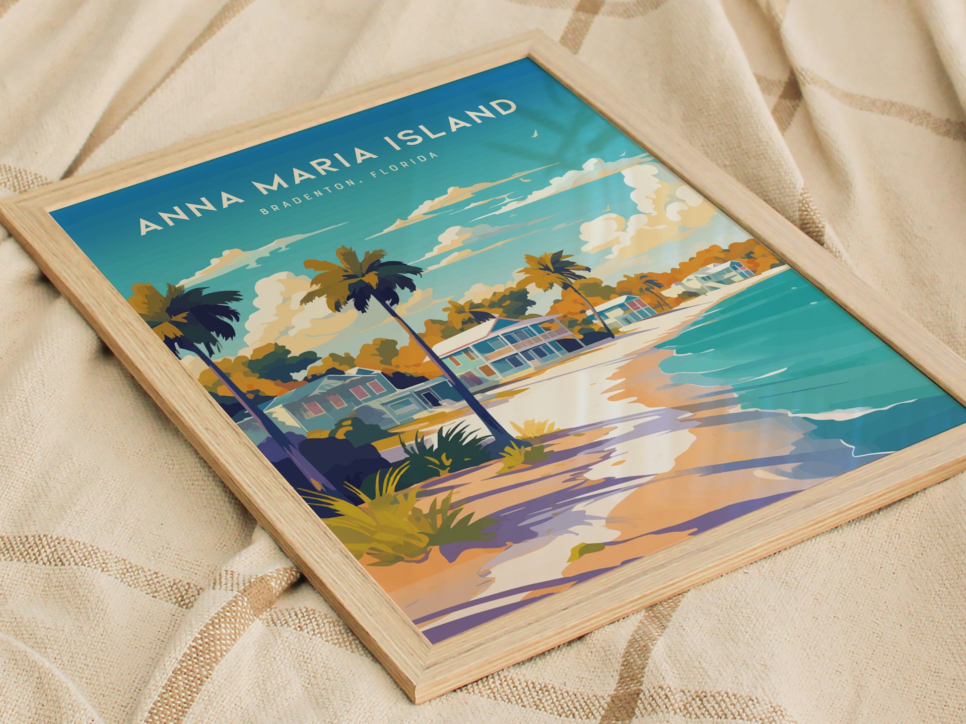 A framed art print of Anna Maria Island, Florida, featuring vibrant illustrations of beach cottages, palm trees, and a serene shoreline. The poster rests on a textured beige fabric, highlighting its coastal theme.