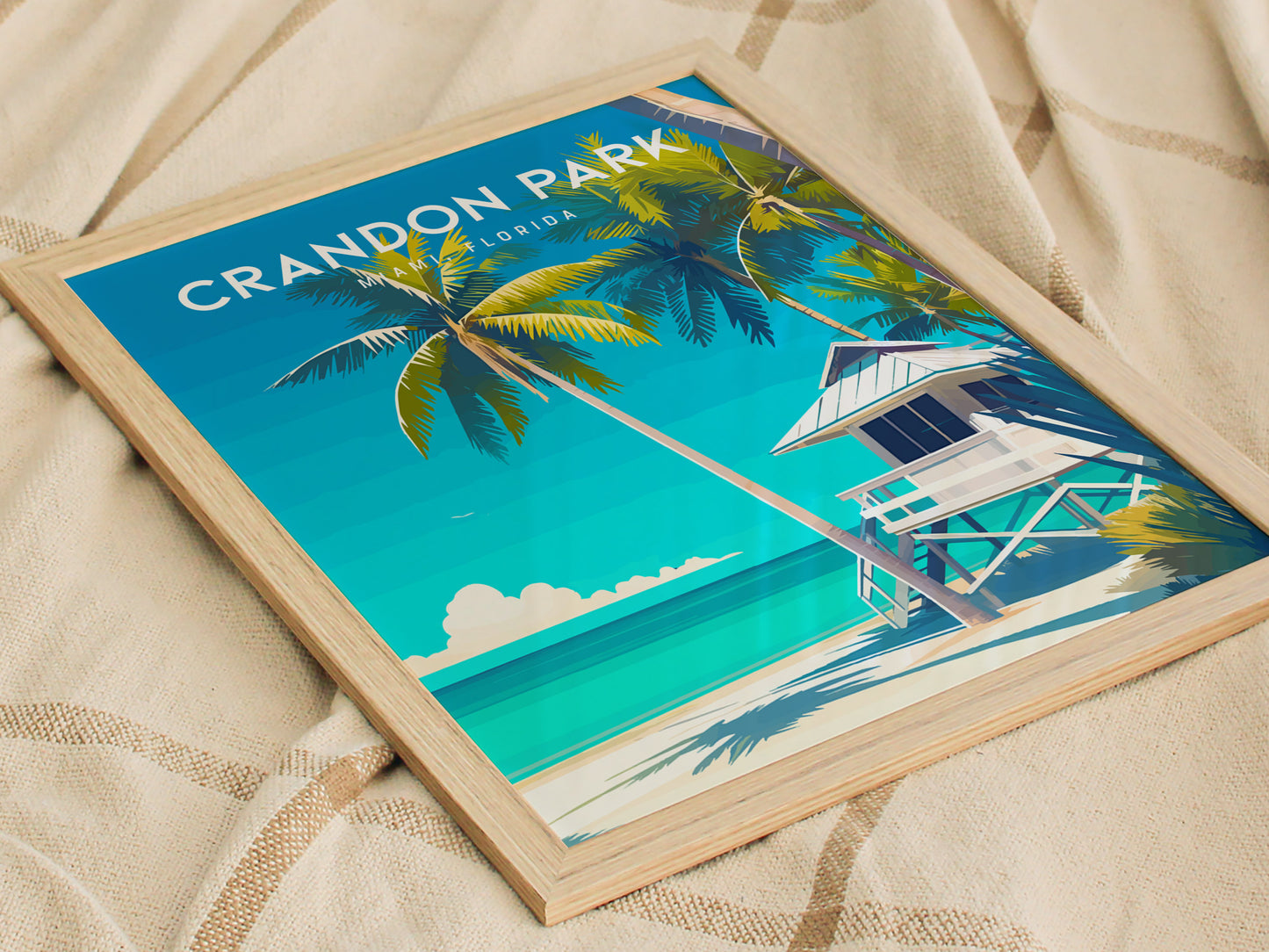 A framed poster of Crandon Park Beach, Miami, features vibrant turquoise waters, a white lifeguard tower, and swaying coconut palms under a clear blue sky. The artwork rests on a beige, textured fabric surface.