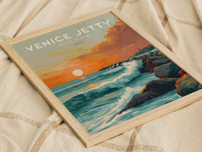 A framed poster titled Venice Jetty depicts a vibrant sunset over waves crashing against a rock jetty, evoking a serene coastal scene. The poster rests on a textured fabric surface.