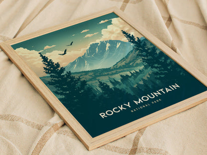 A framed poster of Rocky Mountain National Park featuring illustrated peaks, alpine forests, and birds in flight, lies on a textured beige fabric.