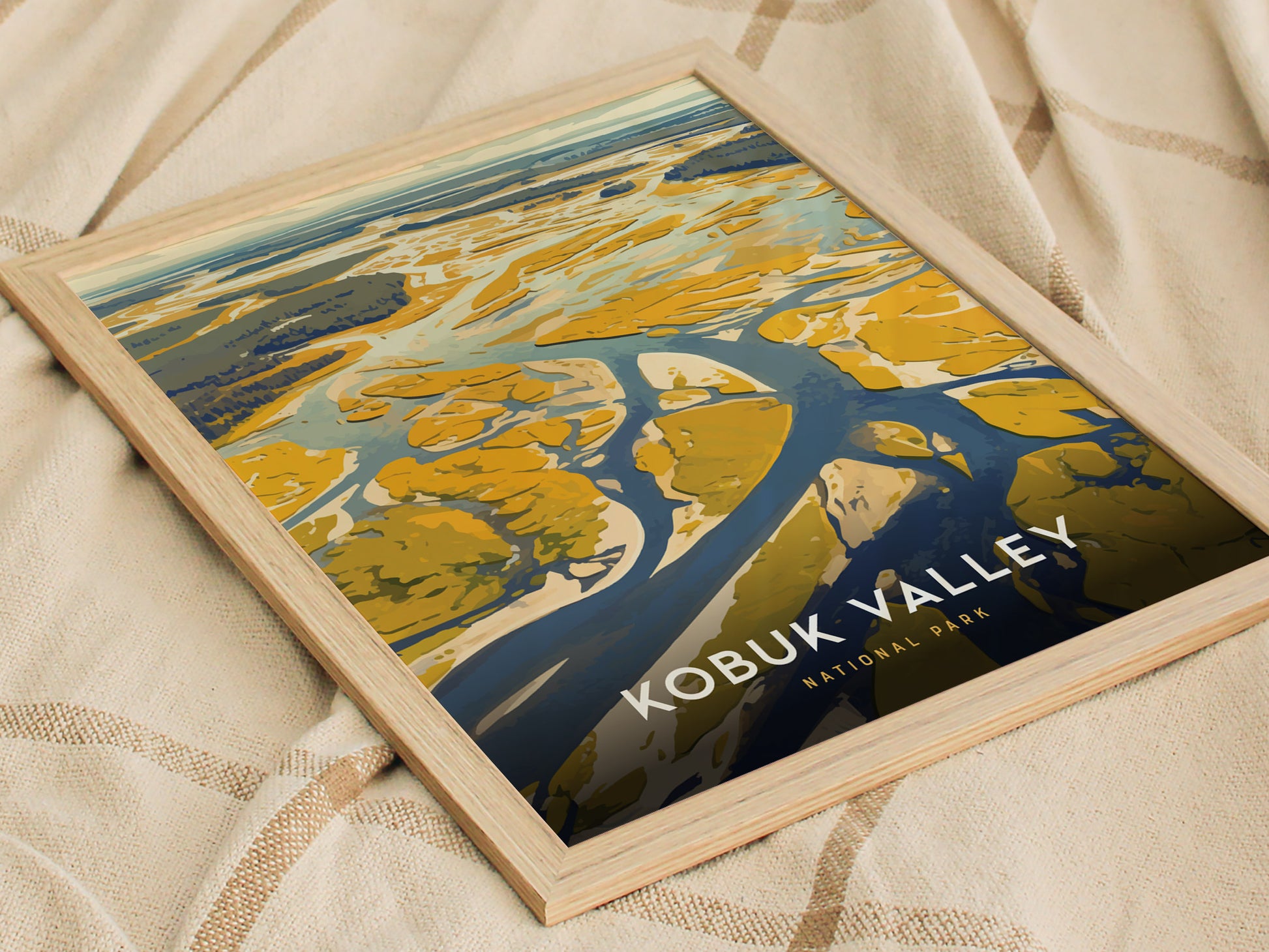 Framed artwork of Kobuk Valley National Park featuring abstract sand dunes and river, placed on a beige fabric. The poster highlights vibrant yellows and blues, with the title visible.