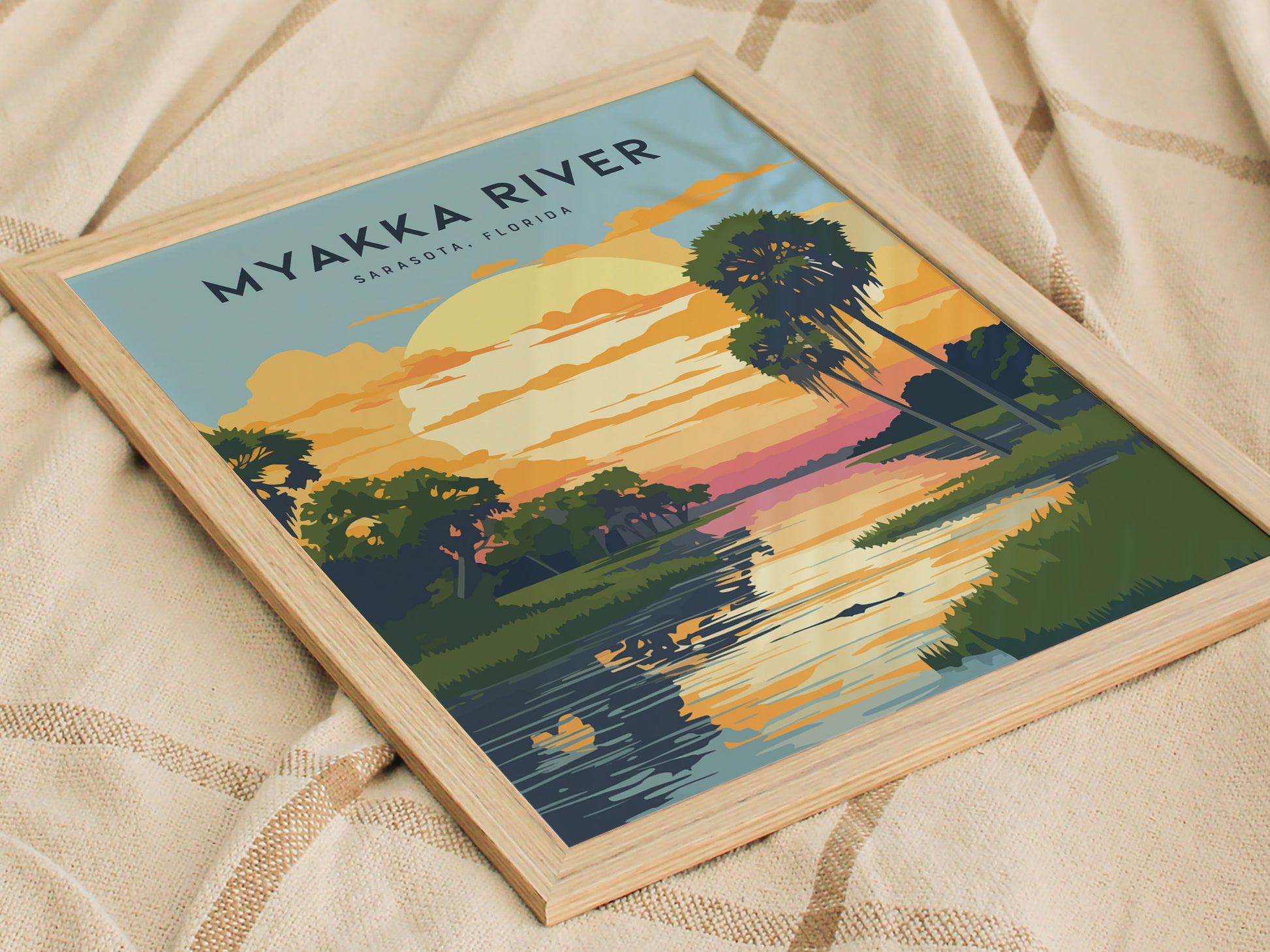 Framed Myakka River State Park poster resting on a textured beige fabric, depicting a vibrant sunset with palm trees and a reflective river scene in Sarasota, Florida.