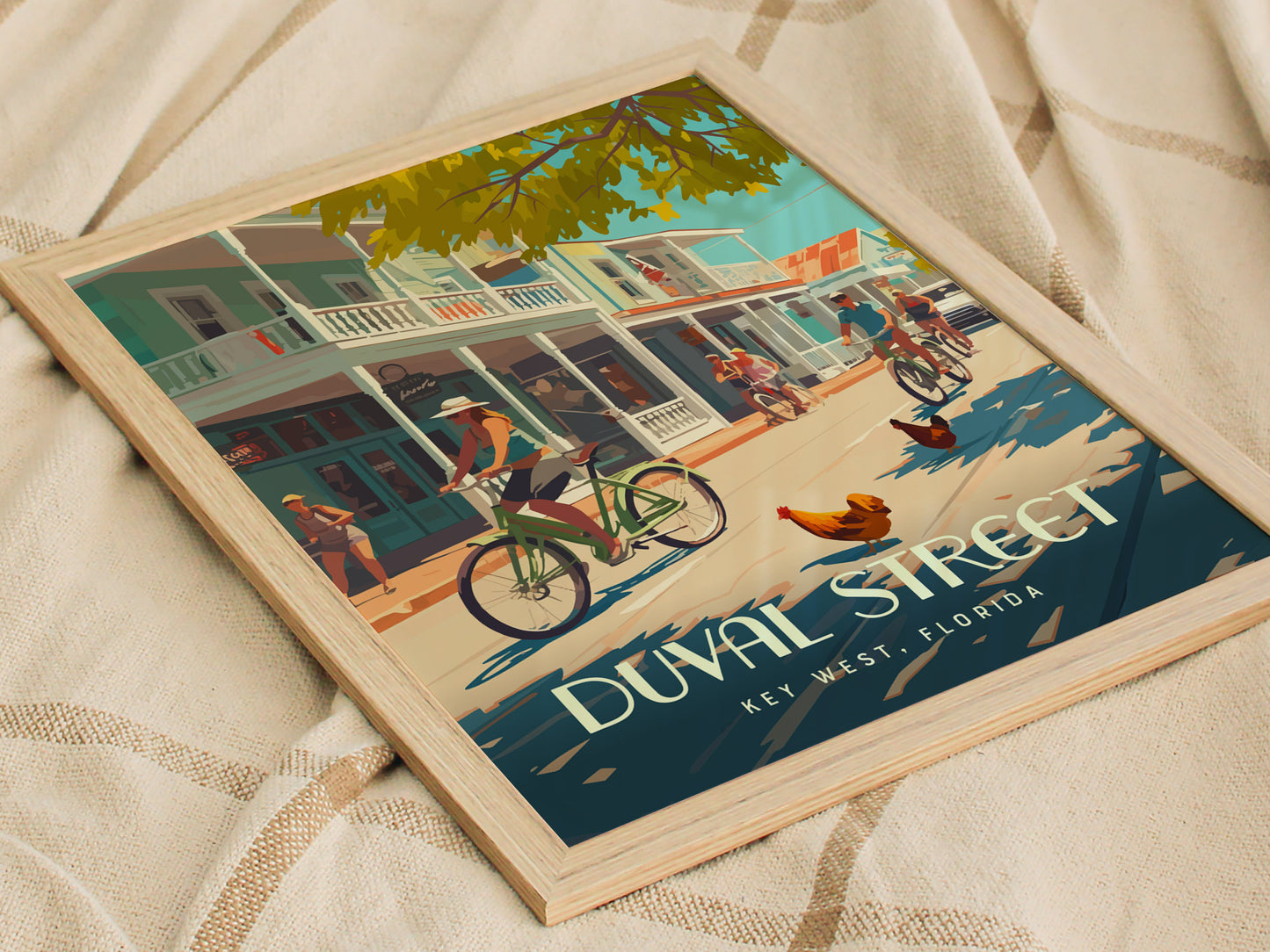 Framed poster depicting a lively scene on Duval Street, Key West, with cyclists, pedestrians, and roosters under a tree-lined street. The poster rests on a textured fabric surface.