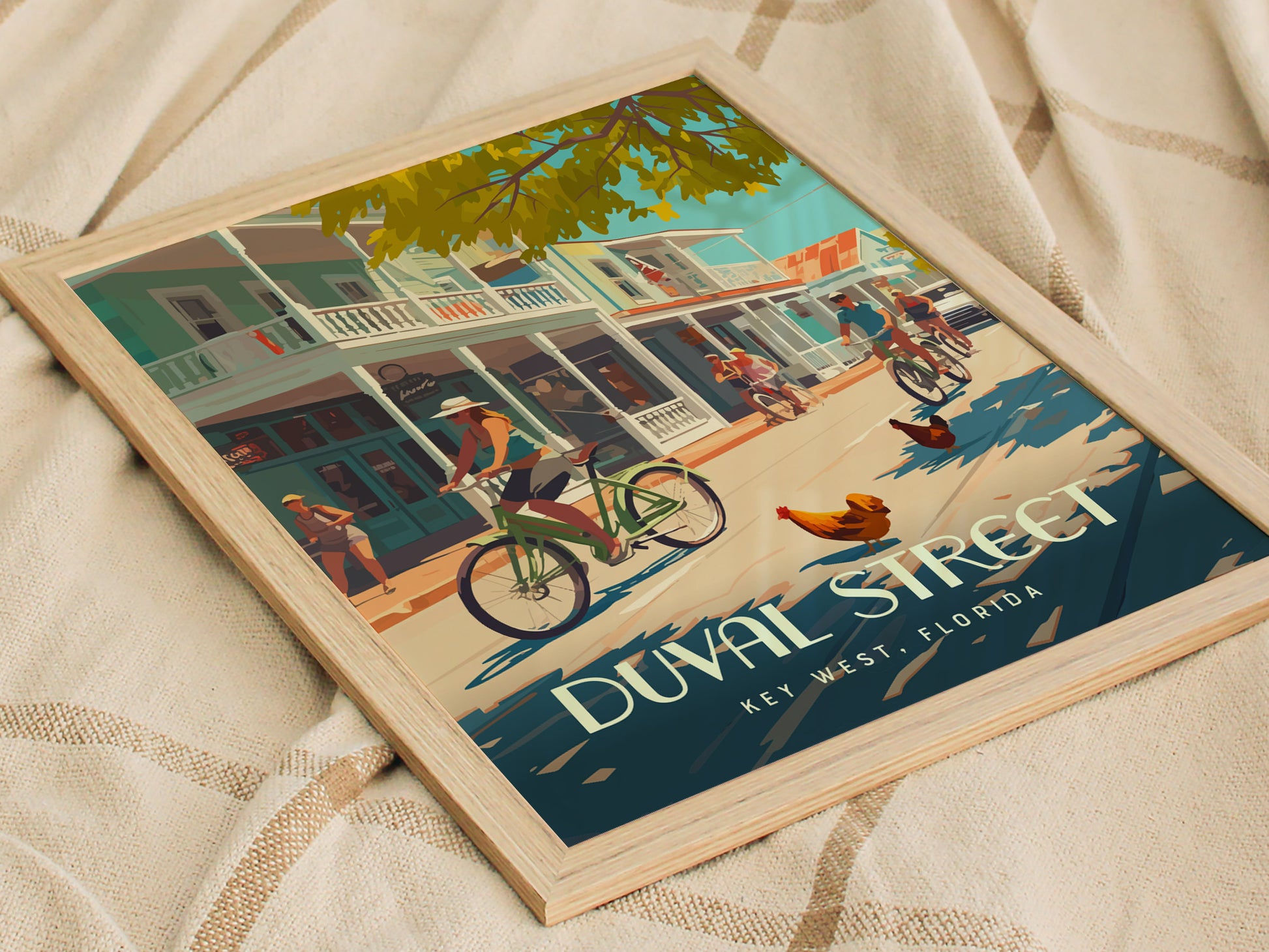 Framed poster depicting a lively scene on Duval Street, Key West, with cyclists, pedestrians, and roosters under a tree-lined street. The poster rests on a textured fabric surface.