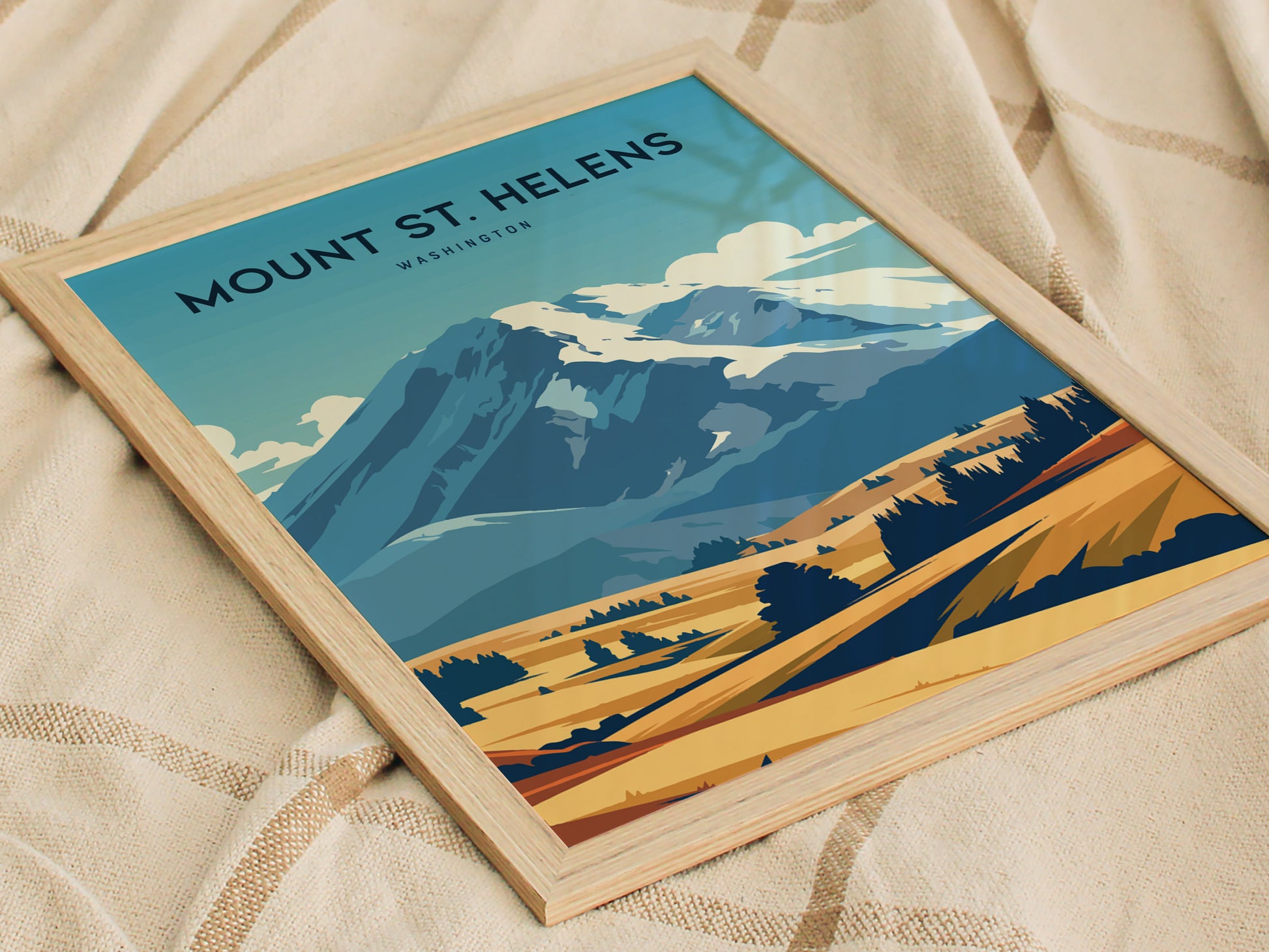 Framed Mount St. Helens poster featuring stylized blue and yellow tones, depicting the mountain and surrounding landscape. The poster rests on a textured beige fabric background.
