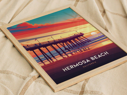 A framed poster of Hermosa Beach, California, features a vibrant sunset over a pier, with vivid orange, pink, and blue hues, resting on a textured fabric surface.