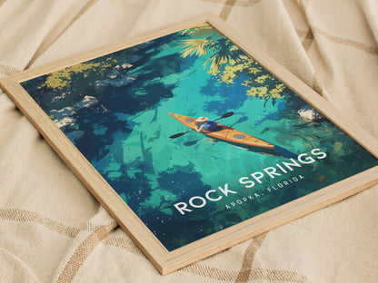 A framed poster displays an illustrated kayaker navigating vibrant blue waters surrounded by lush greenery, with text reading Rock Springs, Apopka Florida. The poster rests on a beige fabric.