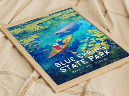 A framed poster featuring two kayakers on vibrant blue water with a manatee swimming nearby, titled Blue Spring State Park, Orange City, Florida, resting on a beige fabric surface.