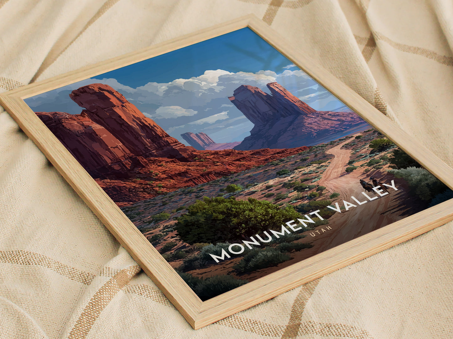 Framed poster of Monument Valley features red sandstone mesas, a winding dirt path, and the iconic Mitten Buttes under a blue sky, placed on a textured fabric surface.