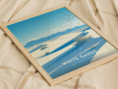 A framed poster of White Sands National Park is placed on a textured beige fabric. The artwork features blue and white sand dunes with distant mountains under a clear sky.