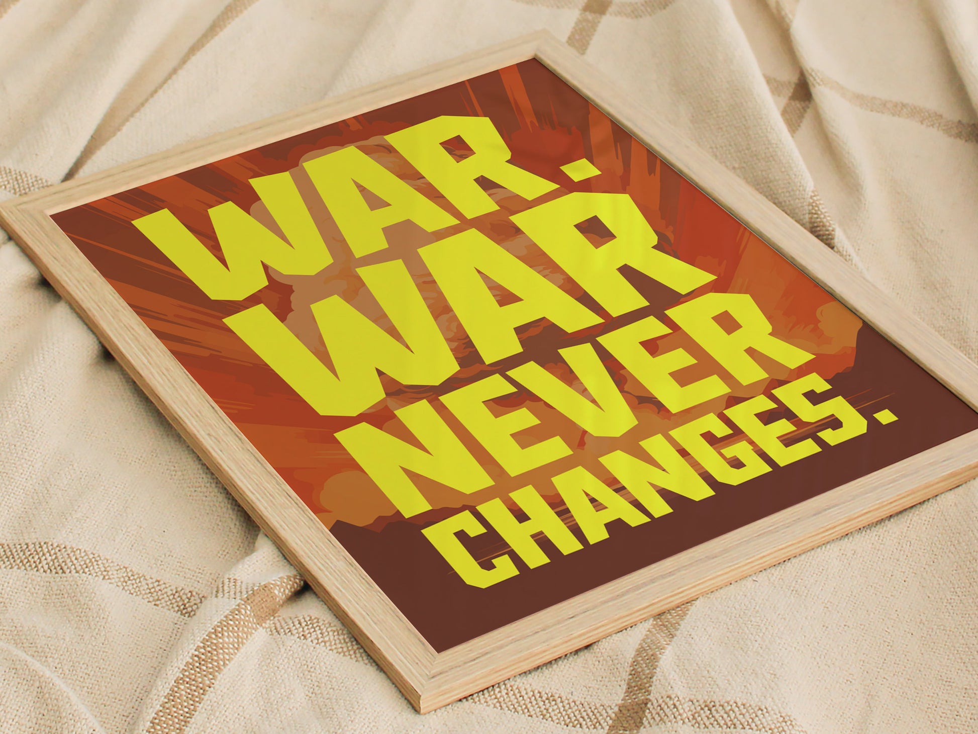 A framed poster with bold yellow text reading War. War Never Changes. against a background of muted reds and oranges, resting on a textured beige fabric.