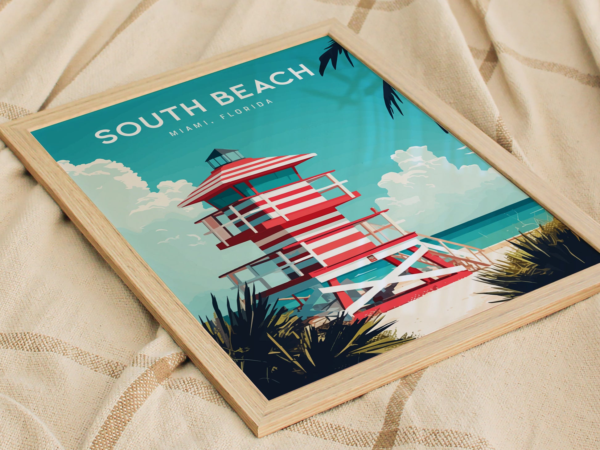 A framed poster of a red and white striped lifeguard tower set against a turquoise ocean and white sandy beach, with South Beach, Miami, Florida written at the top. The artwork is placed on a beige fabric background.