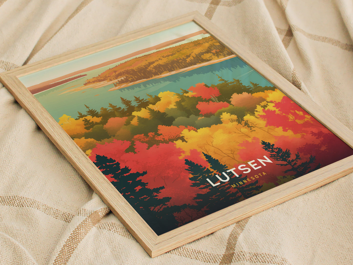 A framed poster featuring colorful autumn foliage with red, orange, and yellow trees against a blue lake and sky, labeled Lutsen Minnesota, rests on a textured beige fabric.