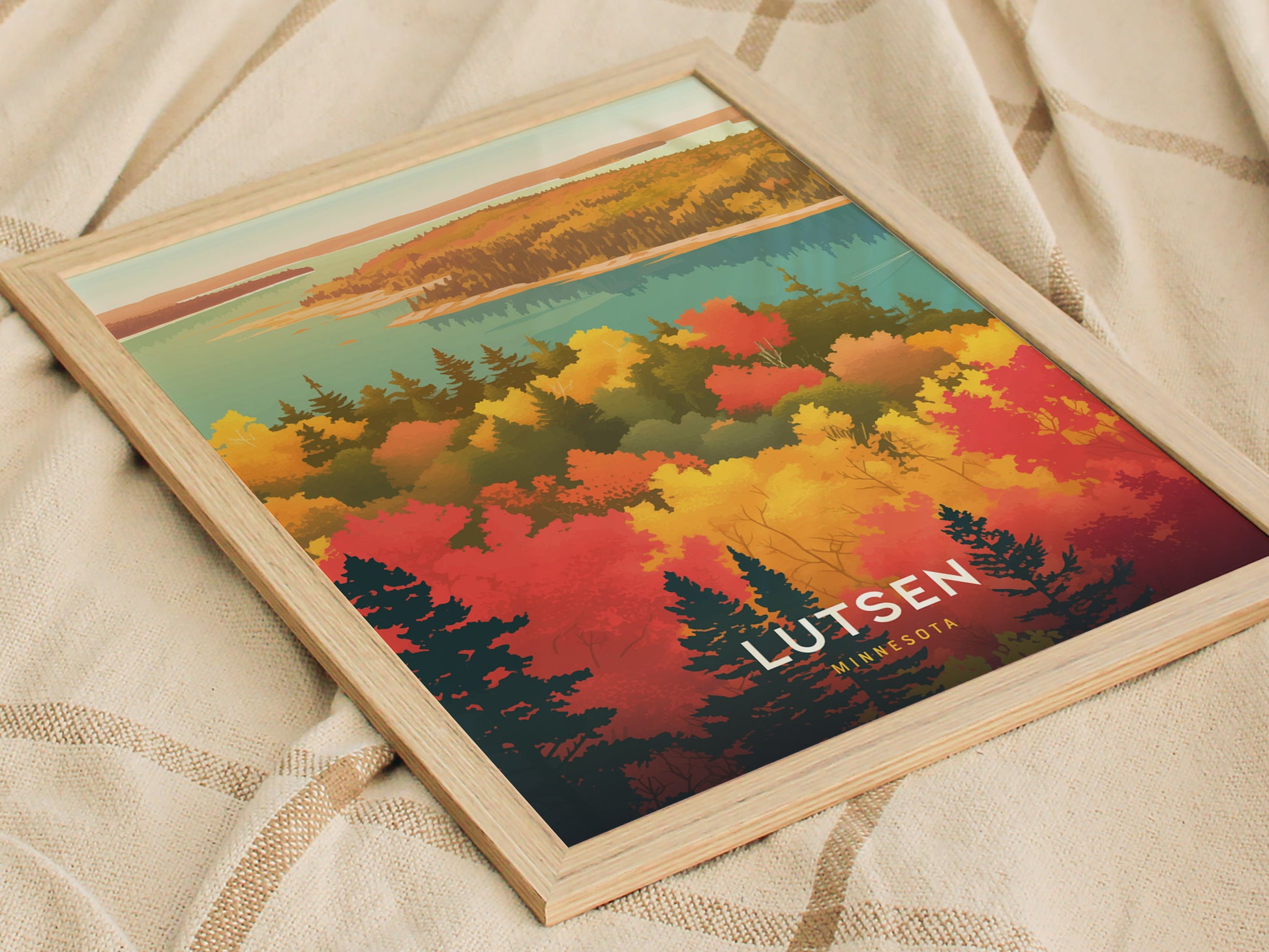 A framed poster featuring colorful autumn foliage with red, orange, and yellow trees against a blue lake and sky, labeled Lutsen Minnesota, rests on a textured beige fabric.