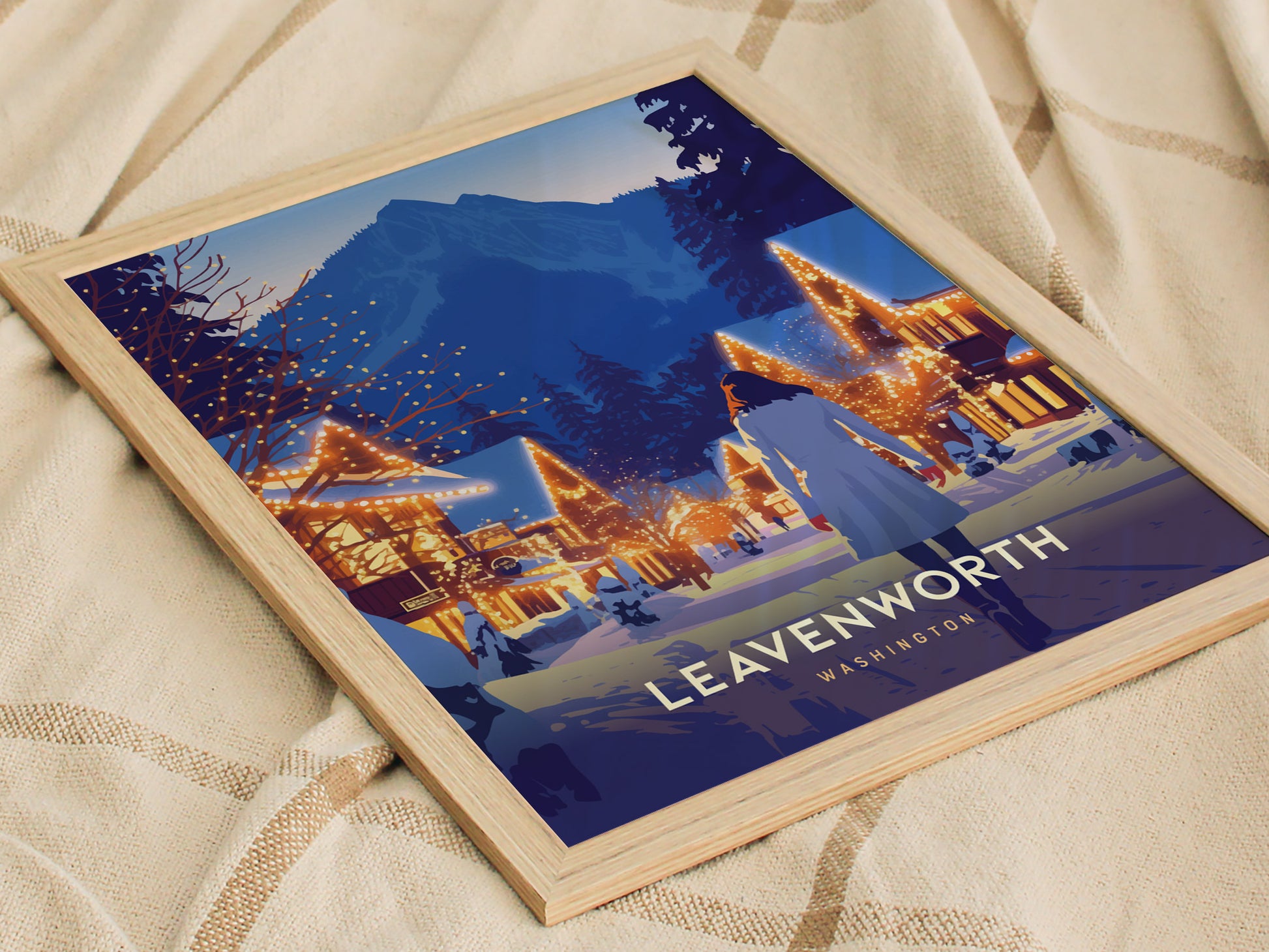 Framed poster of Leavenworth, Washington, featuring a snowy village scene with illuminated buildings against a mountainous backdrop, resting on a patterned fabric surface.