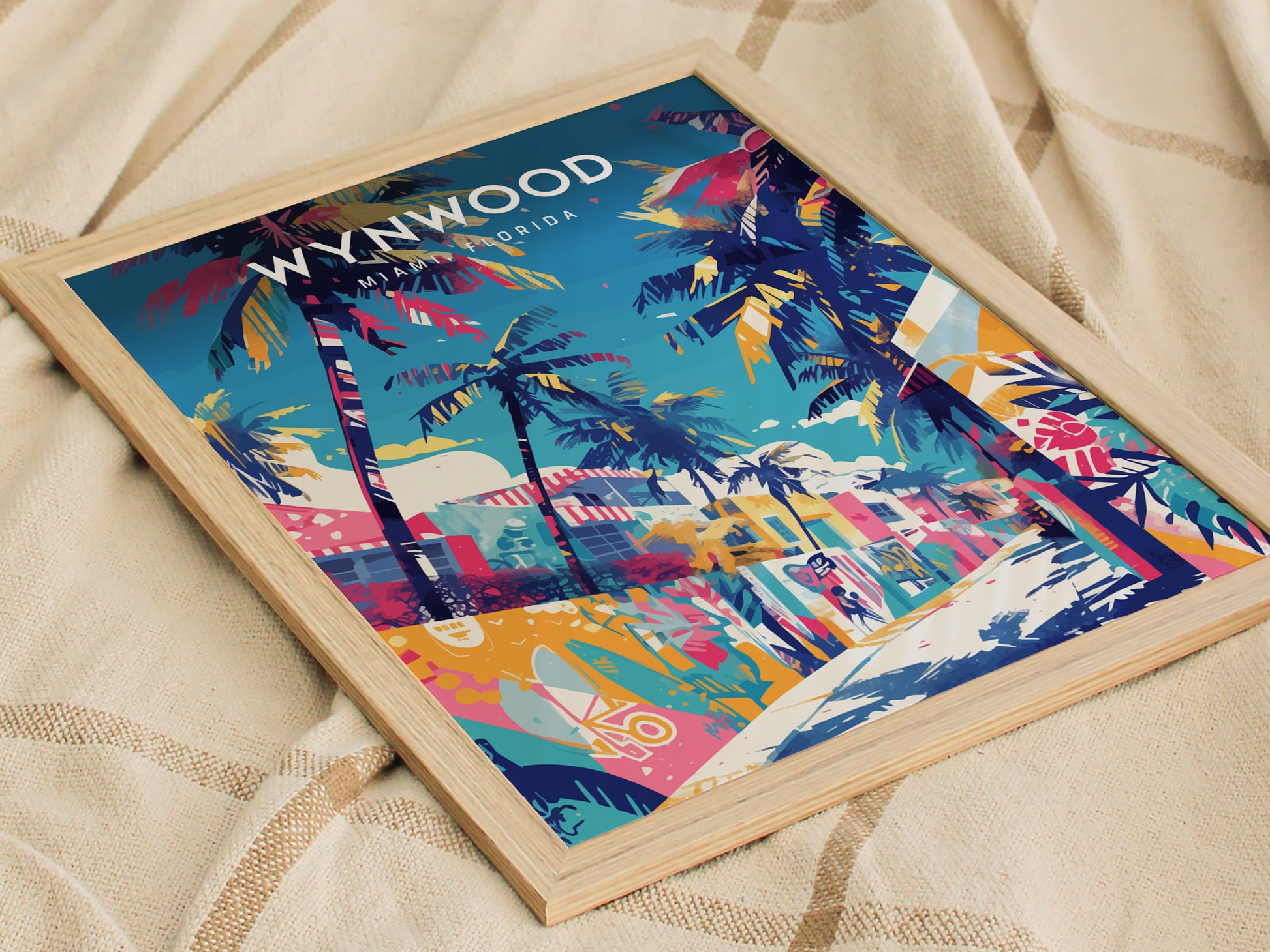 A colorful poster depicting Miamis Wynwood area features vibrant graffiti murals, palm trees, and abstract shapes. The artwork is framed in light wood and lies on a textured beige fabric backdrop.