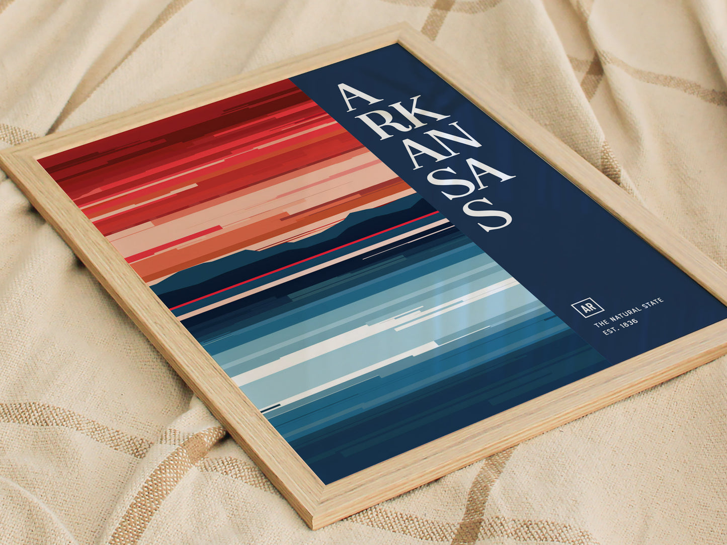 A framed Arkansas poster features a minimalist design with bold, horizontal stripes in warm and cool tones, resting on a beige fabric background.