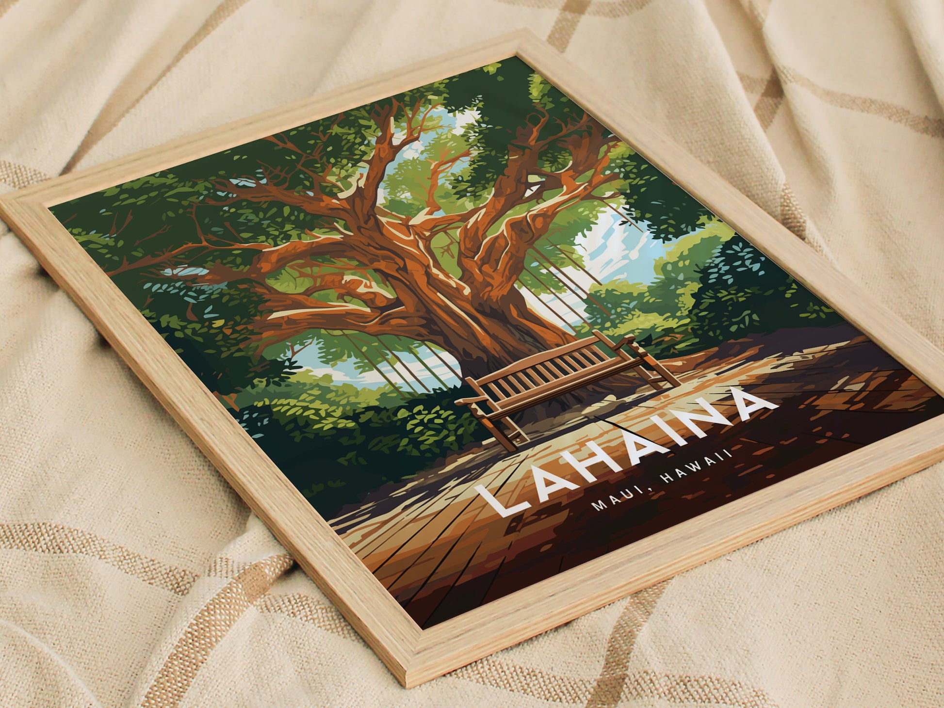 Framed poster of a large banyan tree with a wooden bench underneath, titled Lahaina, Maui, Hawaii, resting on a beige fabric. The artwork features lush greenery and a sunny atmosphere.