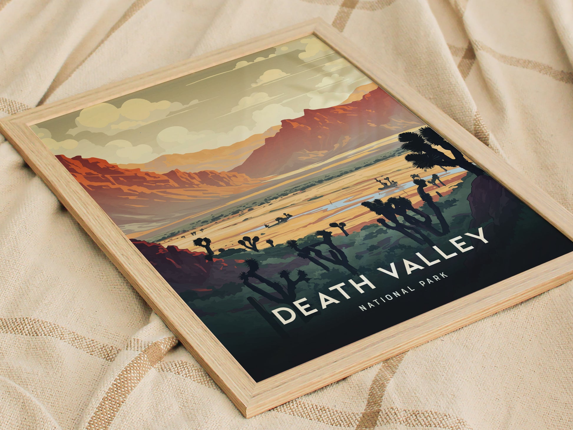 Framed art print depicting Death Valley National Park with vibrant desert landscape, featuring red mountains, cloudy sky, and silhouetted Joshua trees, resting on a textured beige fabric.