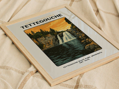 Framed poster featuring an illustration of a waterfall surrounded by cliffs and trees, labeled Tettegouche State Park, Minnesota Est. 1979, resting on a beige fabric surface.