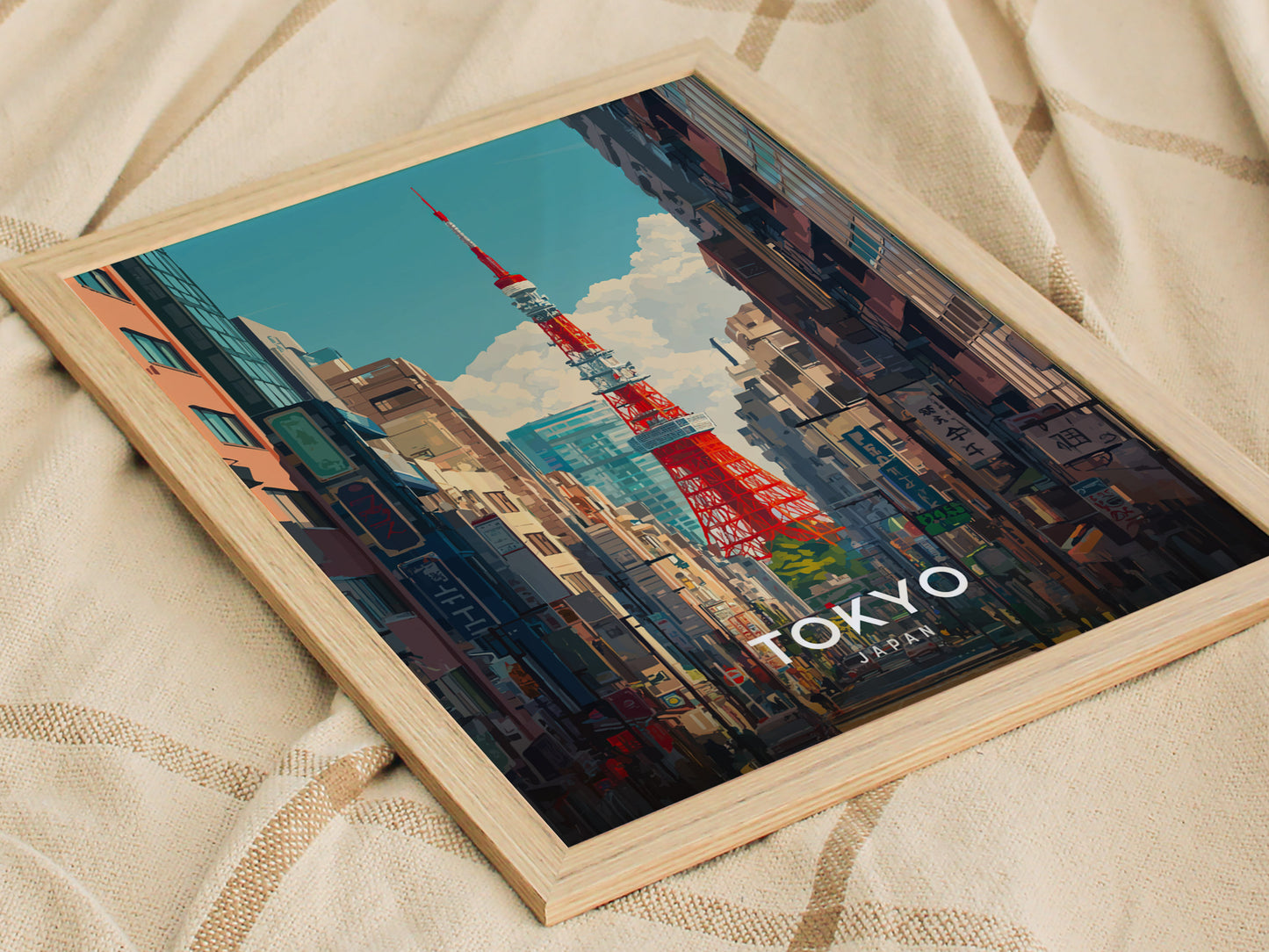 Framed artwork depicting Tokyo Tower rising above a vibrant cityscape, with colorful buildings and clear skies, resting on a textured fabric surface.