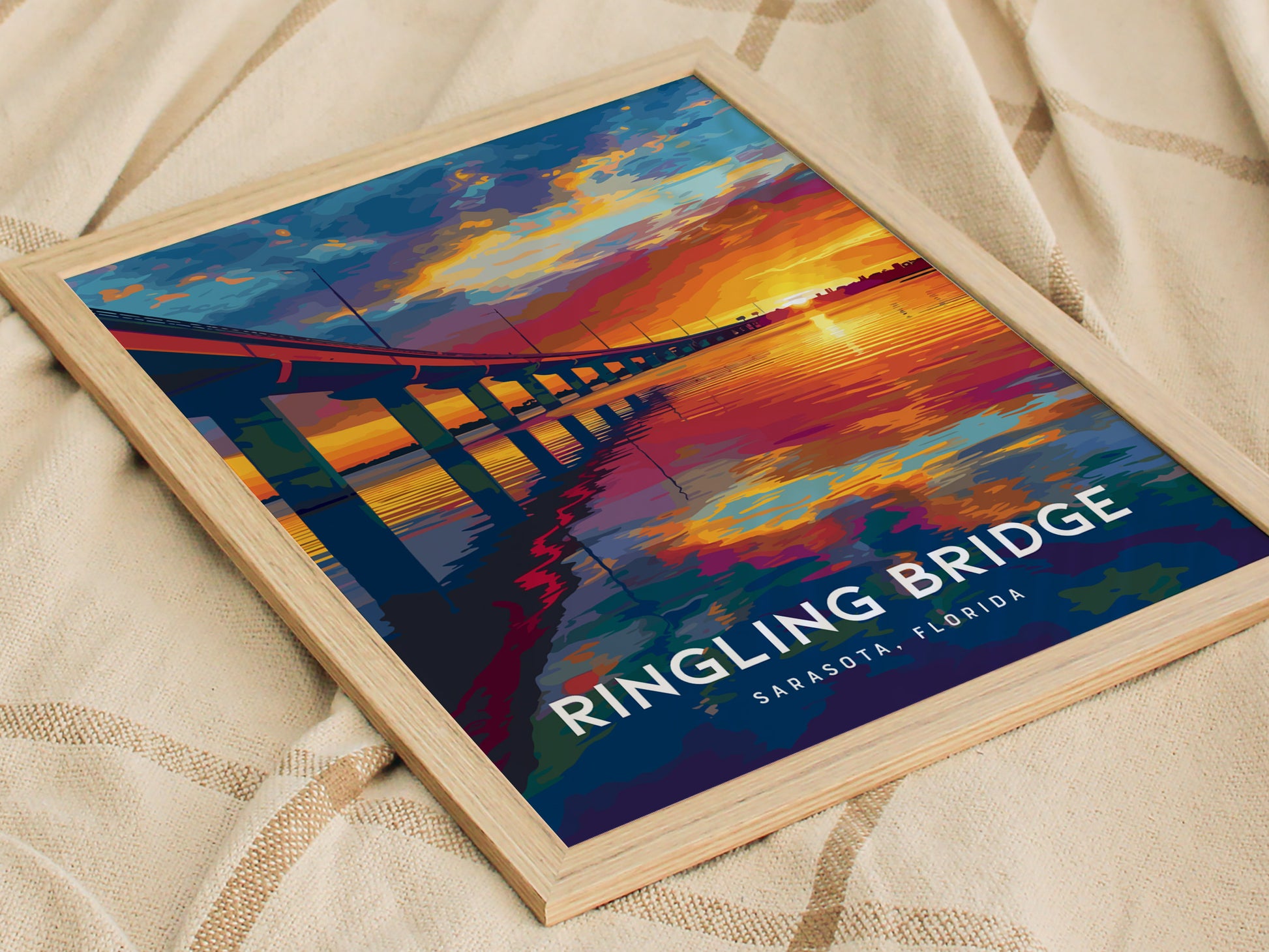 A framed poster of the Ringling Bridge in Sarasota, Florida lies on a beige fabric. The artwork features a vibrant sunset over a body of water.