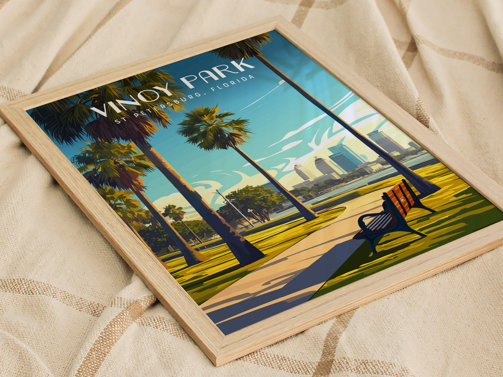 Framed Vinoy Park poster depicting a scenic pathway lined with palm trees, a bench, and distant city skyline under a vibrant blue sky, resting on a textured beige fabric.
